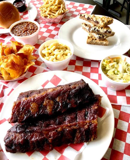 A spread of barbecue ribs, sides of fries, baked beans, macaroni, coleslaw, and dessert on a checkered tablecloth.