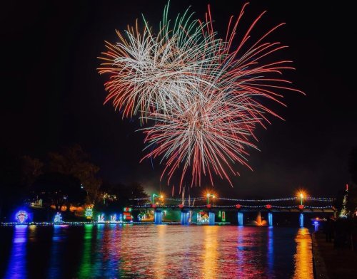 Colorful fireworks illuminate the night sky over a river, with festive lights reflecting on the water.