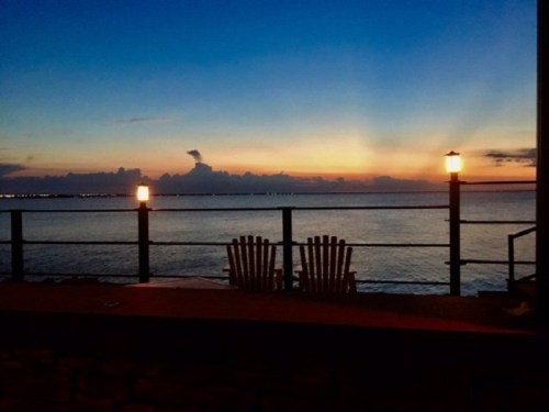 A serene sunset over the water, with two chairs and lanterns along a railing.