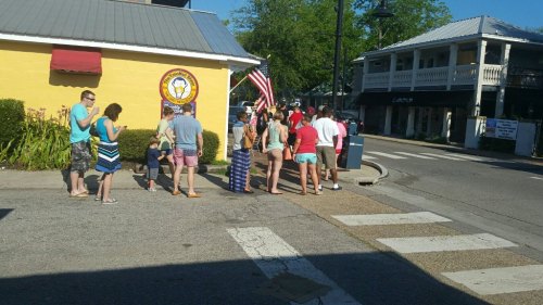 A group of people in casual summer attire waiting in line outside a yellow building with an American flag nearby.