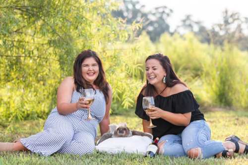 Two women sit on the grass, laughing and holding glasses of wine, with a relaxed animal lying between them.