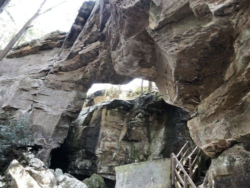 A natural rock arch surrounded by trees and rugged cliffs, with a wooden staircase leading into the area.