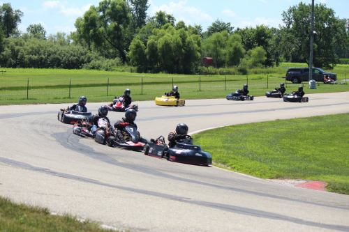 Go-karts racing on a track, with several drivers navigating a curve in a green, outdoor setting.