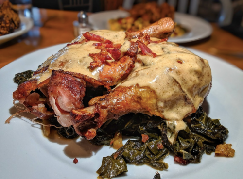 A plate featuring crispy chicken leg topped with creamy sauce and bacon, served on a bed of sautéed greens.
