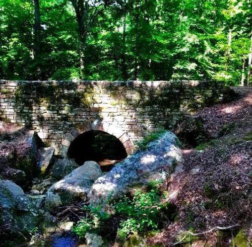 A stone bridge arches over a small stream, surrounded by lush green trees and rocky terrain.