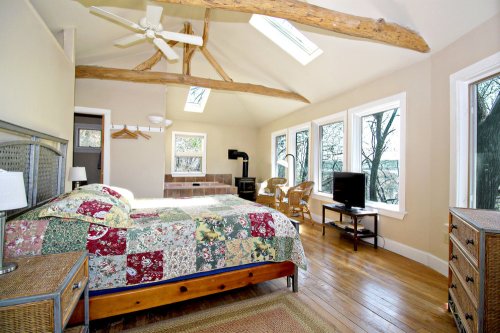 Bright bedroom with a quilted bed, wooden floors, large windows, and rustic beams, featuring a TV and seating area.