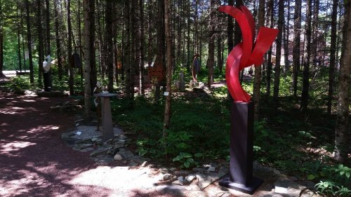 A forest path featuring various sculptures among trees, with a prominent red abstract sculpture in the foreground.
