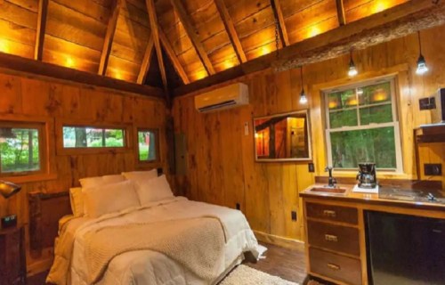 Cozy wooden cabin bedroom with a bed, kitchenette, and warm lighting, surrounded by nature.