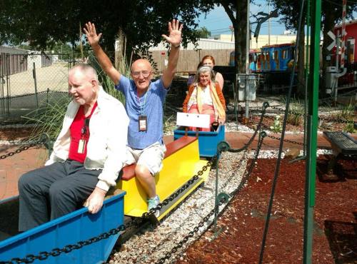 A group of four seniors enjoy a colorful miniature train ride, smiling and raising their hands in excitement.