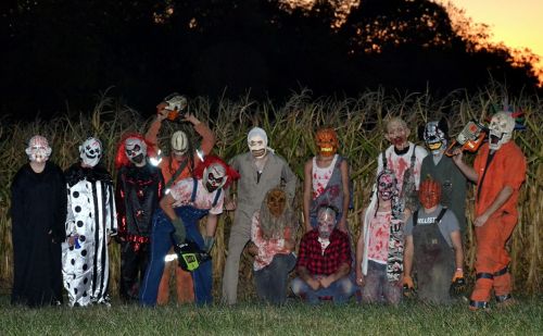 A group of people in costumes and masks, posing in a cornfield at dusk, with a spooky, Halloween theme.