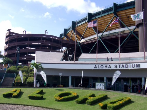 Aloha Stadium entrance with "ALOHA" in flower beds, flags flying, and a parking structure in the background.