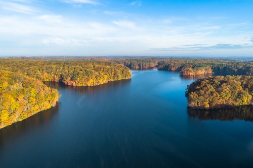 Aerial view of a serene lake surrounded by vibrant autumn foliage and clear blue skies.