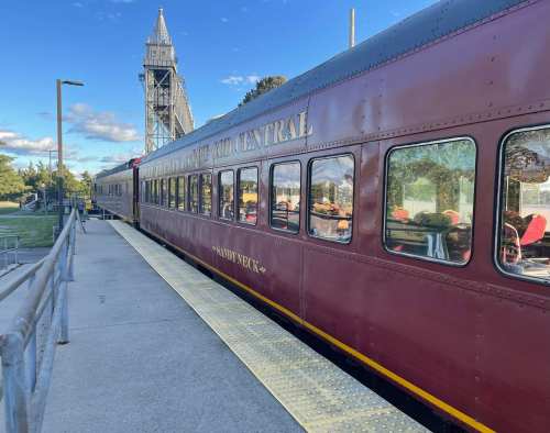 A vintage train car named "Sandy Neck" parked at a station, with a tower and blue sky in the background.