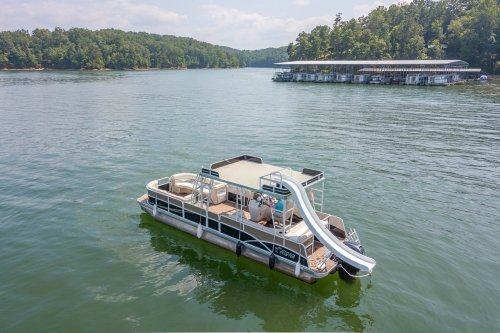 A spacious pontoon boat with a slide, floating on calm water near a dock surrounded by trees.