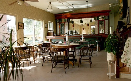 Cozy café interior with wooden tables, chairs, and a bar, featuring large windows and plants for a welcoming atmosphere.
