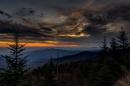 A dramatic sunset over layered mountains, with dark clouds and silhouetted evergreen trees in the foreground.