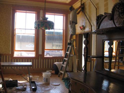 A person on a ladder removes wallpaper in a room with vintage decor and large windows, with painting supplies nearby.
