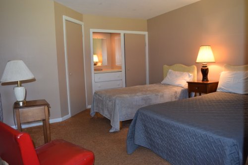 A cozy bedroom with two beds, a lamp, a nightstand, and a mirror reflecting a dresser. Warm, neutral tones.