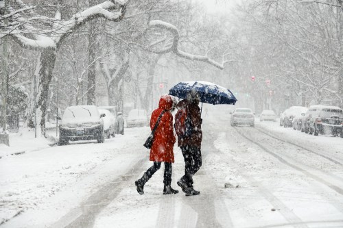 Two people walk in a snowy street, one holding a blue umbrella, while snow falls heavily around them.