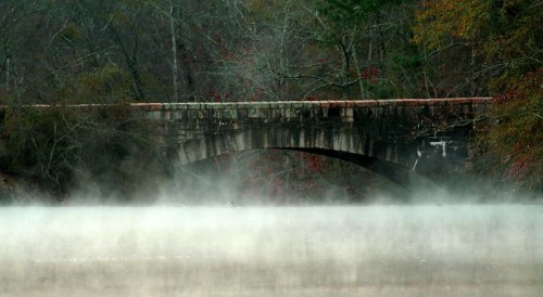 A stone bridge arches over a misty river, surrounded by trees with autumn foliage.