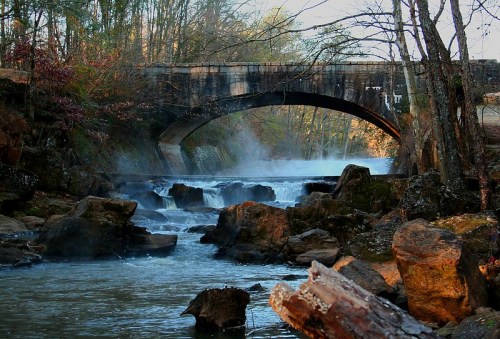 A serene river scene with a stone bridge, mist rising over rocky waters, and trees lining the banks.