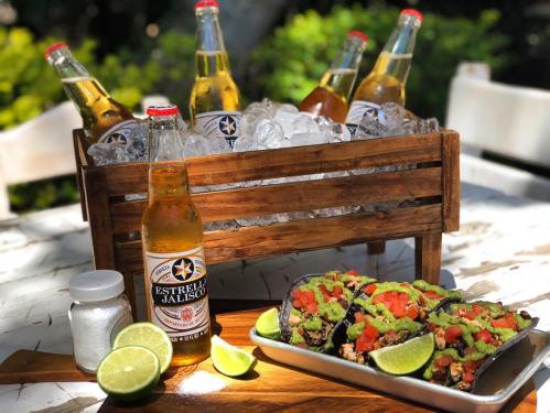 A wooden crate filled with ice and beer bottles, alongside tacos topped with guacamole, tomatoes, and lime wedges.