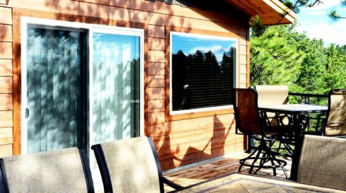 A wooden deck with outdoor furniture, featuring a sliding glass door and large windows surrounded by trees.