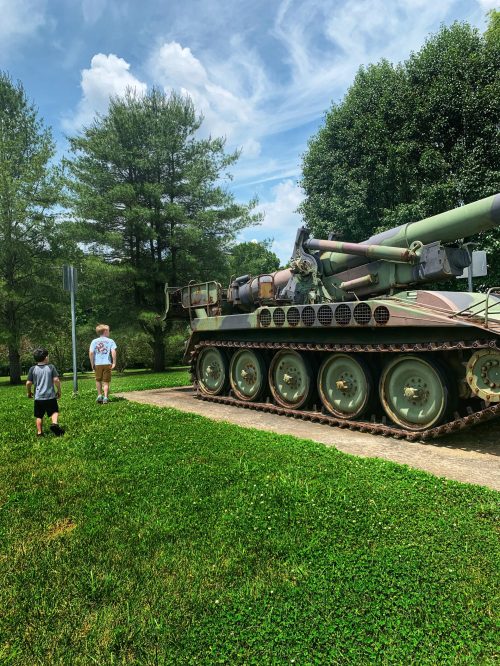 Two children walk past a large military tank on a grassy path, surrounded by trees and a blue sky.
