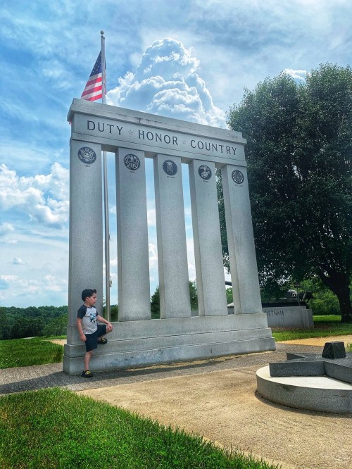A young boy stands beside a tall monument with the words "Duty, Honor, Country" and an American flag against a cloudy sky.