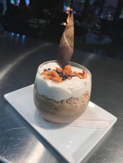 A dessert in a glass featuring layers of cream, chocolate, and topped with pretzels and a decorative leaf.