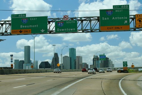 Highway signs directing to Beaumont, Downtown, and San Antonio, with a city skyline in the background and blue skies.