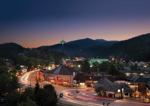 A scenic view of a town at dusk, with mountains in the background and lights illuminating the streets and buildings.