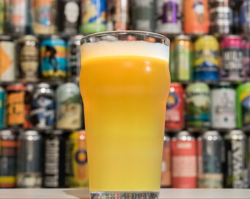 A glass of hazy yellow beer sits in front of a colorful wall of various beer cans.