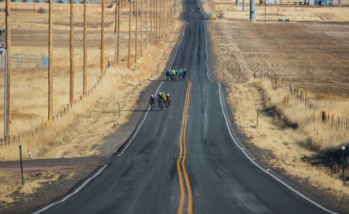 A group of cyclists rides along a long, empty road surrounded by dry fields and power lines under a clear sky.