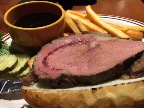 A plate featuring a slice of prime rib on bread, served with fries, pickles, and a small bowl of sauce.