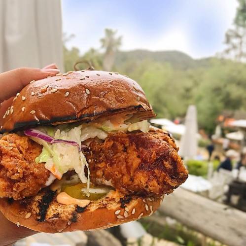 A hand holding a sesame seed bun filled with crispy fried chicken, lettuce, and pickles, with a blurred outdoor background.