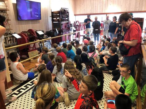 A group of children sitting on the floor in a museum, watching a presentation near old printing equipment.