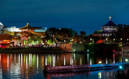 Night view of a serene lake reflecting illuminated traditional buildings and trees, with a clear sky above.