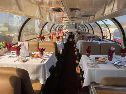 Interior of a dining car with tables set for a meal, featuring white tablecloths and red napkins, overlooking a scenic view.