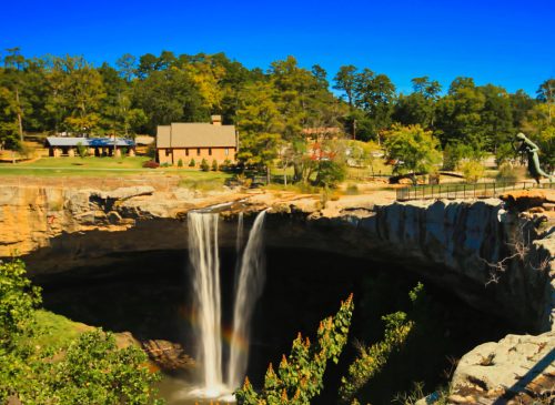 A scenic view of a waterfall cascading into a rocky gorge, surrounded by lush greenery and a distant building.