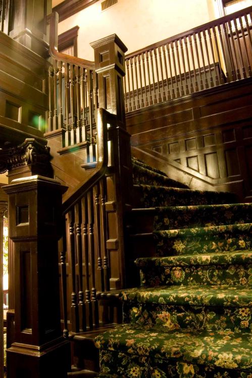 A wooden staircase with ornate railings and a floral carpet, leading to an upper level in a vintage-style interior.