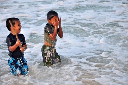 Two children play in the ocean waves, one laughing and the other covering his face as water splashes around them.