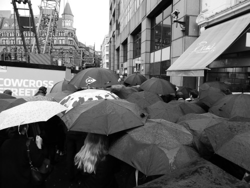 A crowd of people with umbrellas in a rainy urban setting, buildings in the background, captured in black and white.