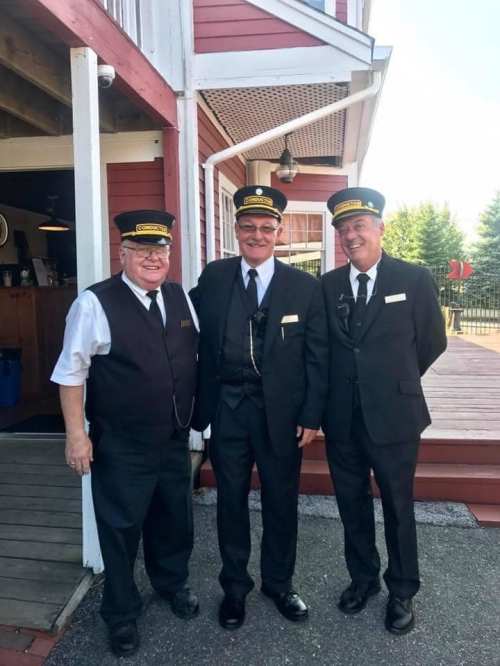 Three train conductors in uniforms and hats stand together outside a building with a red exterior.