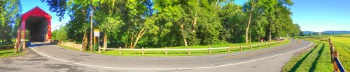 A scenic view of a red covered bridge beside a winding road, surrounded by lush greenery and fields.