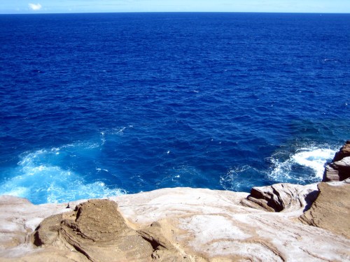 A rocky shoreline meets a deep blue ocean under a clear sky, with gentle waves lapping at the rocks.