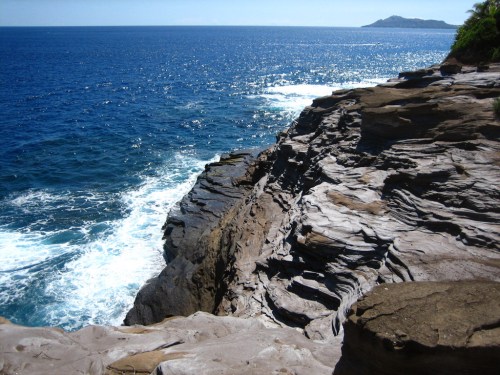 Rocky coastline with waves crashing against the shore, overlooking a sparkling blue ocean and distant island.