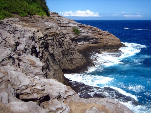 Rocky coastline with waves crashing against the shore, under a clear blue sky and distant boat on the water.