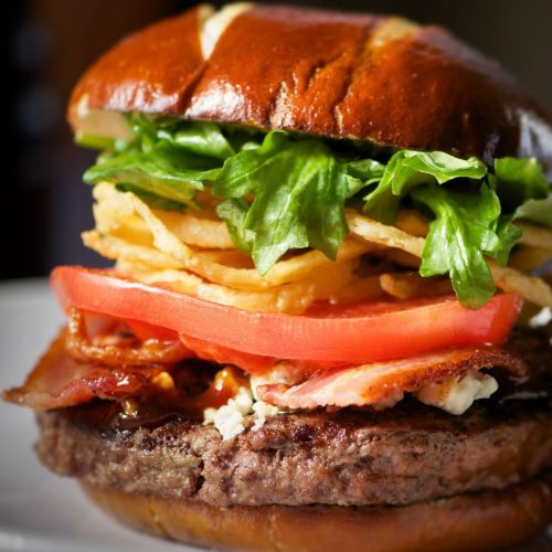 A close-up of a gourmet burger with a pretzel bun, lettuce, tomato, bacon, and crispy fries stacked inside.