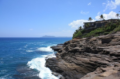 Coastal view with rocky cliffs, ocean waves, and a house surrounded by palm trees under a clear blue sky.